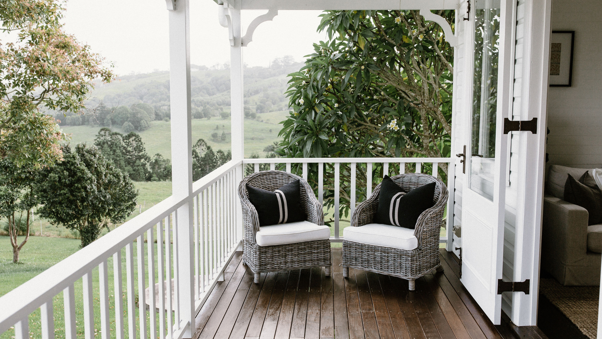 Two wicker chairs on a wooden verandah featuring white French doors and a backdrop of beautiful country scenery.